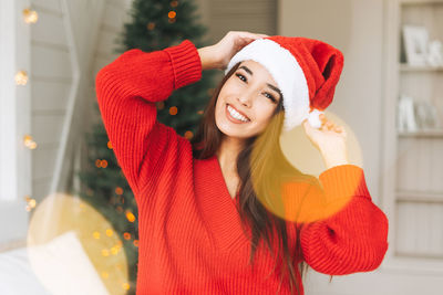Young asian woman in red knitted sweater and santa hat having fun on bed in room with christmas tree