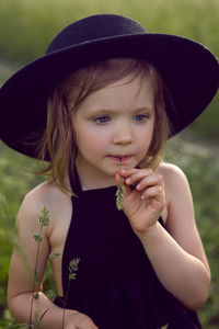 Child girl in a black hat and dress stands in a field with a grass in her mouth in summer