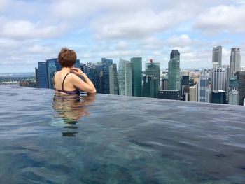Woman looking at modern building while standing in infinity pool against cloudy sky