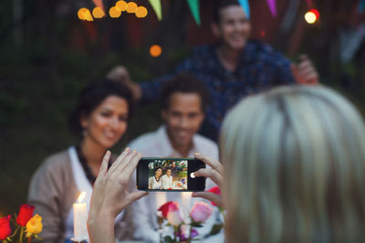 Cropped image of woman photographing happy friends through smart phone at garden party