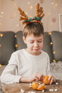 Boy in christmas antlers, sits and peels a tangerine on the background of a yellow garland.