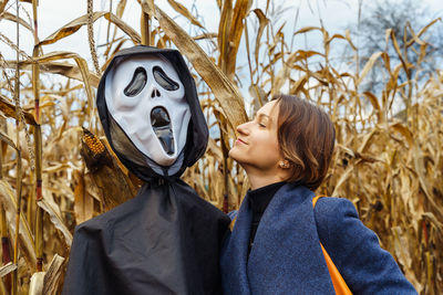 Young woman next to figure with death mask in the middle of a cornfield on halloween