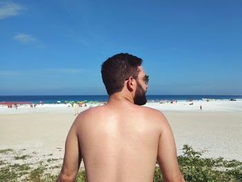 Rear view of shirtless man standing at beach against blue sky