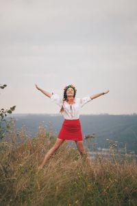 Smiling young woman with arms raised standing on field against clear sky