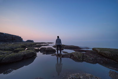 Rear view of man standing on rocks at shore against sky