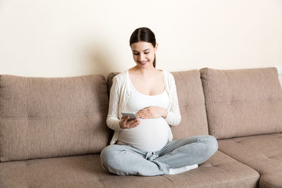 Caucasian pregnant girl using cellphone, resting on bed in home.