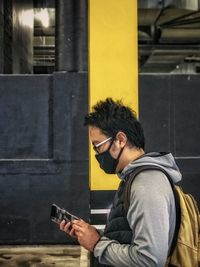 Side view of young man using mobile phone against yellow pillar.