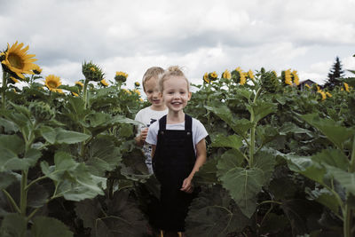 Portrait of sister with brother walking amidst sunflowers against cloudy sky at farm