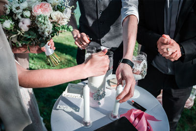 Midsection of bride and groom holding candles on table by friend