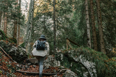 Rear view of woman wearing backpack and hat, hiking on path in moody green forest.