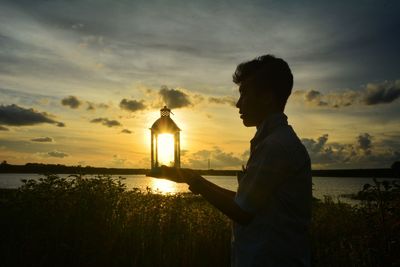 Side view of silhouette man holding lantern by sea at sunset