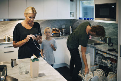 Boy standing by mother using mobile phone and father cleaning plates in dishwasher at kitchen