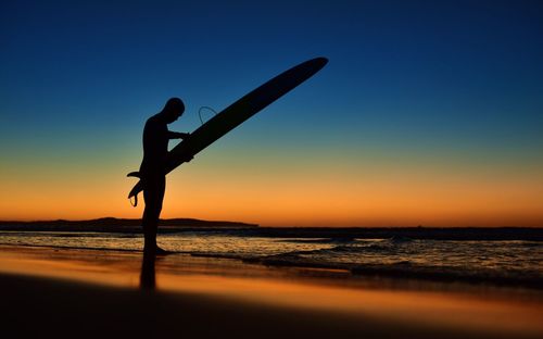 Silhouette of man holding surfboard at beach during sunset