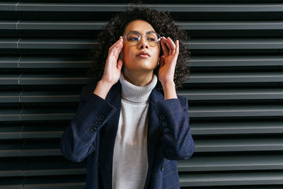 Serious self assured young ethnic female in stylish clothes touching eyeglasses and looking away while standing against gray striped wall