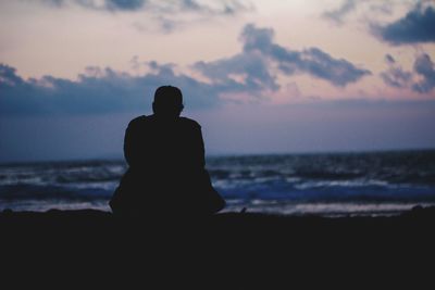 Silhouette man sitting on beach against sky during sunset