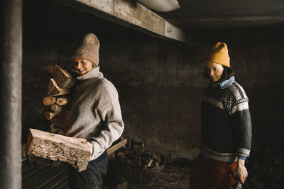 Mature woman looking at girlfriend collecting firewood while standing in shed