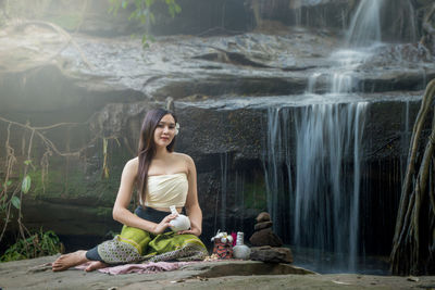 Portrait of smiling young woman sitting on rock against waterfall