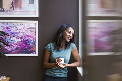 Thoughtful woman holding coffee cup while standing against picture frames on wall at home
