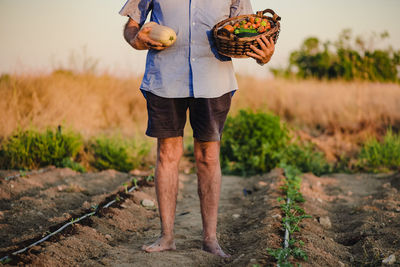 Cropped unrecognizable aged male farmer standing barefoot with wicker basket of fresh fruits and vegetables on garden bed and looking away