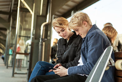 Young man showing mobile phone to teenage girl while sitting on railroad station platform