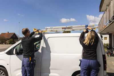 Rear view of male and female electricians getting ladder from van on sunny day