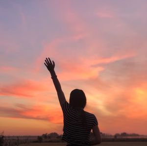 Rear view of woman waving hand while standing against sky during sunset