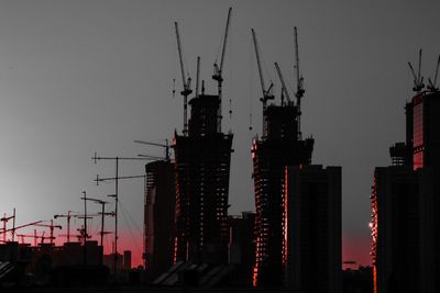 Silhouette cranes by buildings against sky during sunset