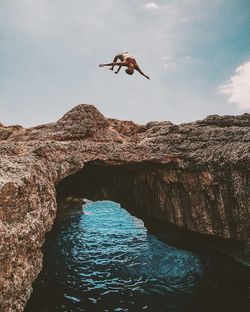 Man jumping on rock formation in sea against sky