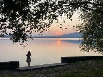 Rear view of man standing by lake against sky during sunset