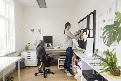 Full length of artist sticking documents on bulletin board in creative office