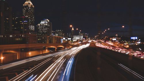 Light trails on road by illuminated buildings against sky at night