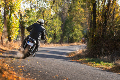 Man in leather jacket and helmet riding bike on asphalt road in sunny autumn day in countryside