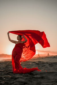 Full length of woman wearing red dress standing in beach against sky during sunset