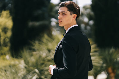 A beautiful young man, the groom in an elegant wedding suit, stands posing in the city's old park