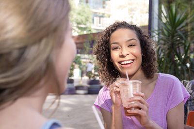 Happy woman with drink looking at friend while sitting at sidewalk cafe