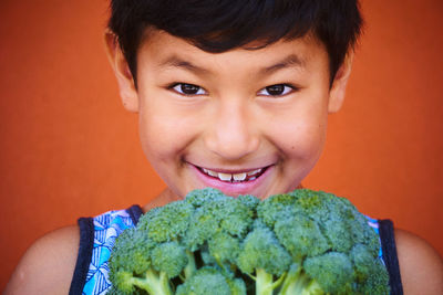 Close-up portrait of smiling boy holding broccoli against orange background