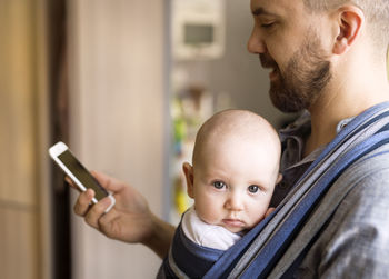 Father with baby son in sling at home using cell phone