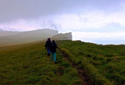 Full length rear view of hikers walking on grassy cliff against cloudy sky
