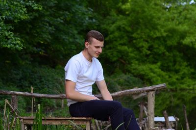 Young man sitting stool by railing against trees