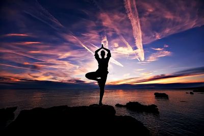 Silhouette person doing yoga on rock formation against sea at sunset