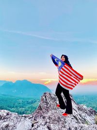 Woman holding malaysian flag while standing mountain against sky during sunset