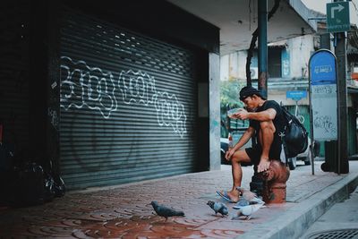 Side view of young man with backpack feeding birds while sitting on bollard in city