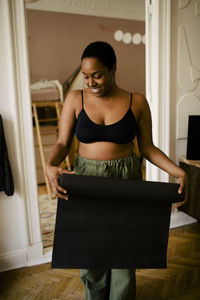 Smiling young woman with exercise mat standing at home