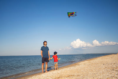 Father and son are standing on a sandy beach by the sea and launch a toy striped kite in the summer