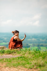 Side view of young woman using smart phone while sitting on mountain against sky