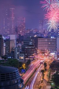 Light trails on city street by buildings against sky at night