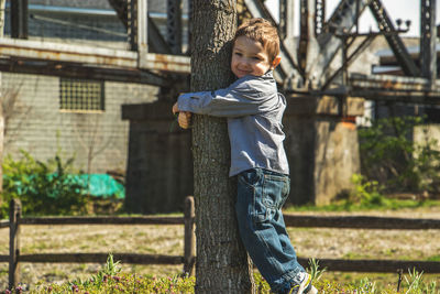 Portrait of cute boy hugging tree against building