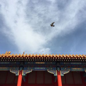 Low angle view of bird flying over traditional building against cloudy sky