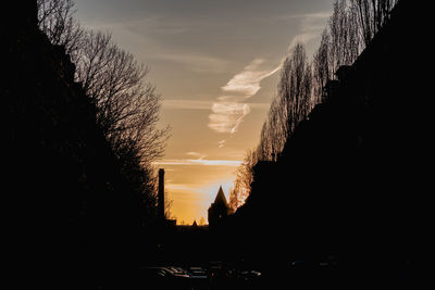 Silhouette trees by buildings against sky during sunset