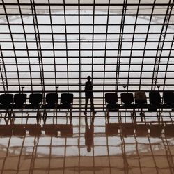 Rear view of man standing at departure area against window with reflection on tiled floor at airport